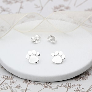 Puede incluir: Un par de pendientes de plata con forma de huella de pata. Los pendientes son pequeños y delicados, y serían un gran regalo para cualquier amante de los perros.