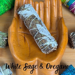 May include: A wooden hand-shaped dish holding a bundle of white sage and oregano tied with white string. The text White Sage & Oregano is printed across the center. Surrounding the dish are various colorful herb bundles in green, rainbow, and blue.