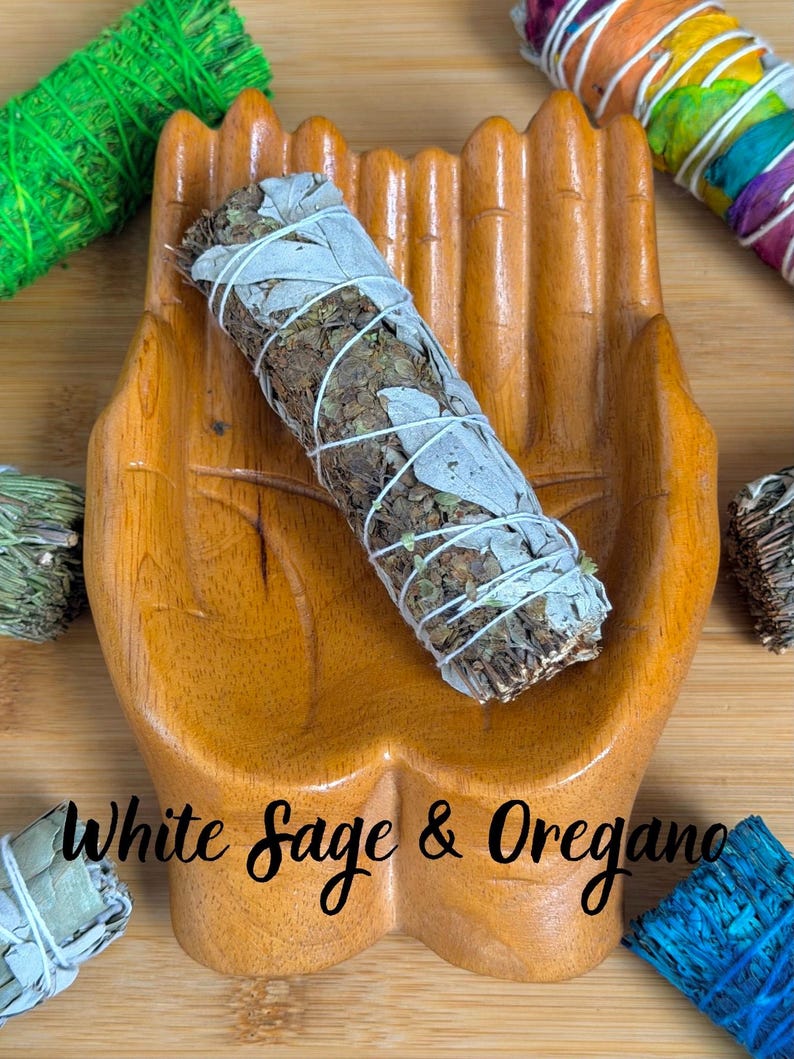 May include: A wooden hand-shaped dish holding a bundle of white sage and oregano tied with white string. The text White Sage & Oregano is printed across the center. Surrounding the dish are various colorful herb bundles in green, rainbow, and blue.