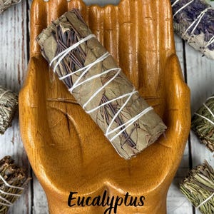 May include: A wooden bowl shaped like two cupped hands holding a bundle of dried eucalyptus leaves wrapped in white string. The word Eucalyptus is printed in black script at the bottom of the bowl, surrounded by other bundles of dried herbs.