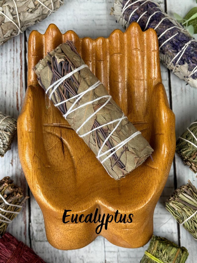 May include: A wooden bowl shaped like two cupped hands holding a bundle of dried eucalyptus leaves wrapped in white string. The word Eucalyptus is printed in black script at the bottom of the bowl, surrounded by other bundles of dried herbs.
