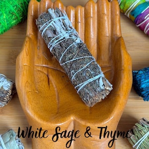 May include: A wooden hand-shaped dish holding a bundle of dried white sage and thyme tied with white string. The text "White Sage & Thyme" is printed on the dish. Other colorful herb bundles are visible in the corners of the image.