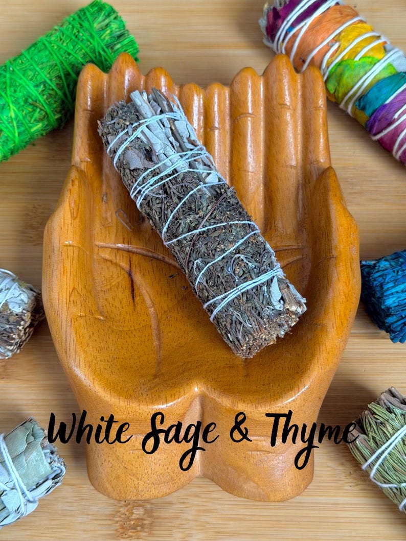 May include: A wooden hand-shaped dish holding a bundle of dried white sage and thyme tied with white string. The text "White Sage & Thyme" is printed on the dish. Other colorful herb bundles are visible in the corners of the image.