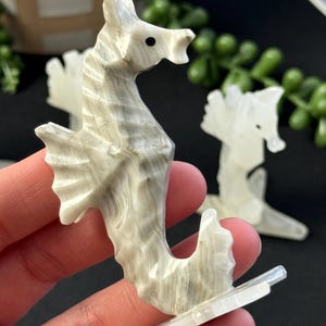 Puede incluir: Una figurita de caballito de mar tallada en blanco y gris con un ojo negro. El caballito de mar est&aacute; de pie sobre una base blanca.