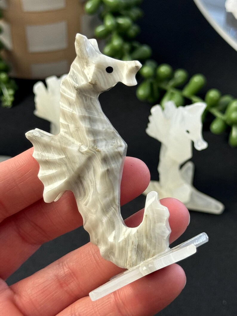 Puede incluir: Una figurita de caballito de mar tallada en blanco y gris con un ojo negro. El caballito de mar est&aacute; de pie sobre una base blanca.
