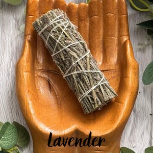 May include: A wooden dish carved in the shape of two cupped hands holding a cylindrical bundle of dried green herbs tied with white string. The word Lavender is printed in black script at the bottom of the wooden hand-shaped dish.