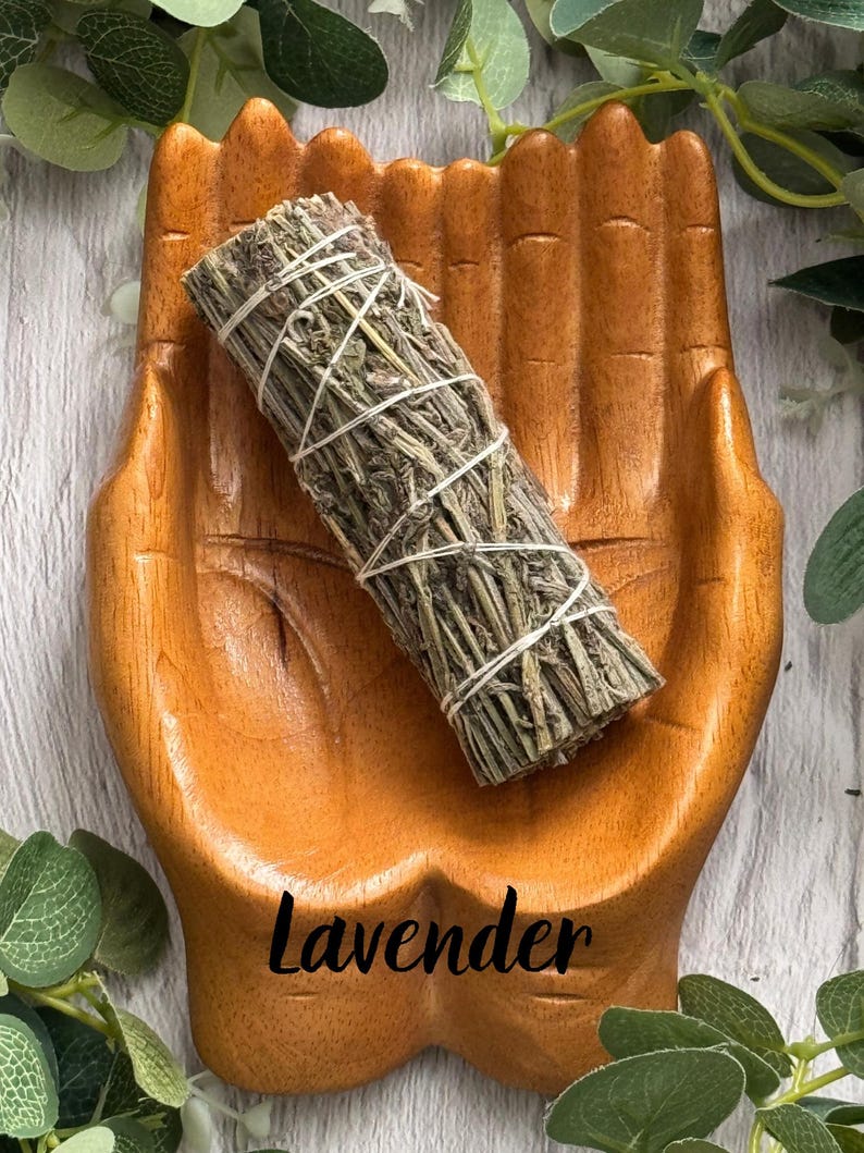 May include: A wooden dish carved in the shape of two cupped hands holding a cylindrical bundle of dried green herbs tied with white string. The word Lavender is printed in black script at the bottom of the wooden hand-shaped dish.