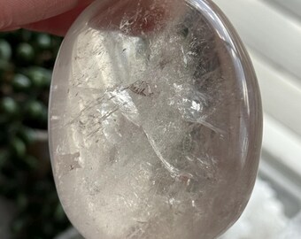 May include: A clear, oval-shaped crystal with internal fractures and inclusions. The polished stone has a translucent appearance, with a light brown hue. The crystal is held in a hand, showcasing its smooth, rounded form.