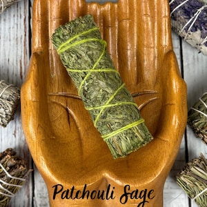 May include: A wooden dish shaped like two cupped hands holding a cylindrical bundle of green dried herbs wrapped in lime green string, with the text Patchouli Sage printed in black script at the bottom of the wooden hand-shaped dish.