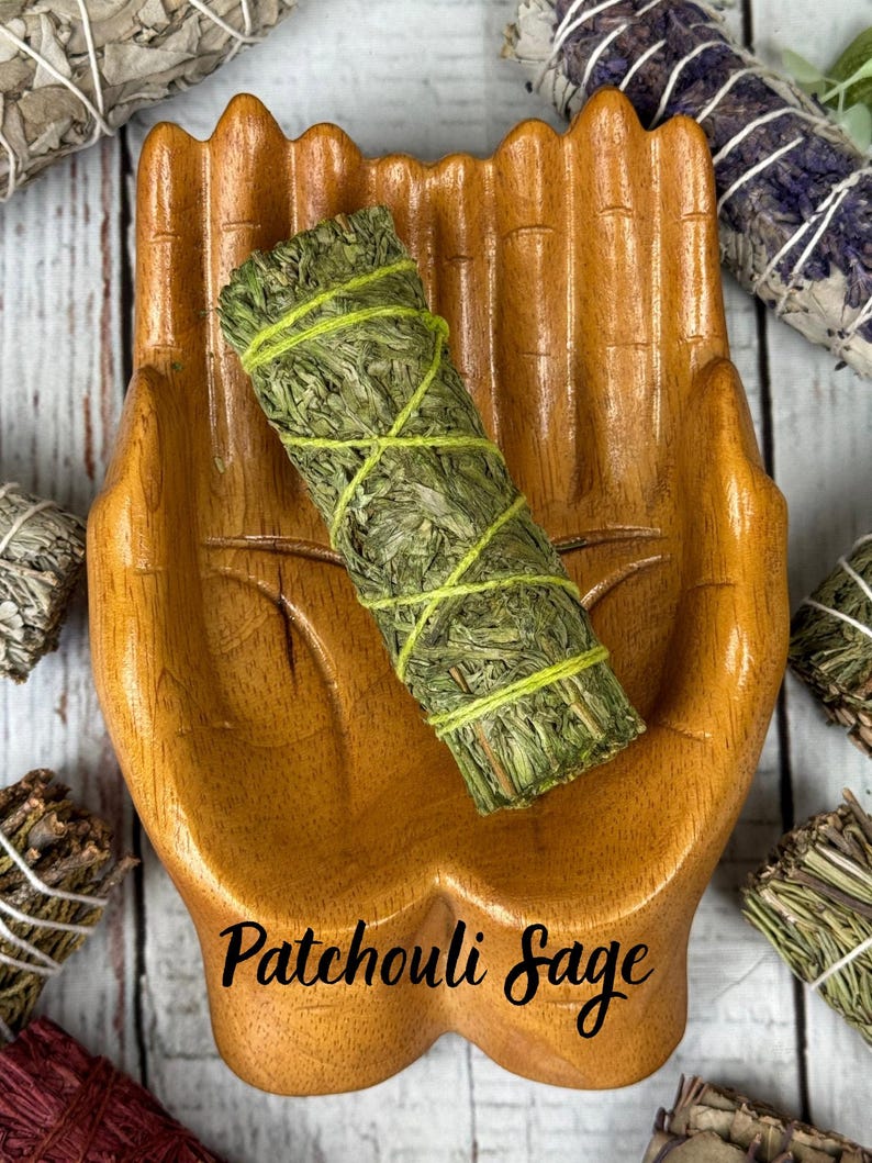 May include: A wooden dish shaped like two cupped hands holding a cylindrical bundle of green dried herbs wrapped in lime green string, with the text Patchouli Sage printed in black script at the bottom of the wooden hand-shaped dish.