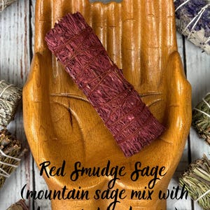 May include: A cylindrical bundle of red smudge sage tied with thin string, resting in a carved wooden hand-shaped dish. The text on the image reads: Red Smudge Sage (mountain sage mix with dragon's blood resin).
