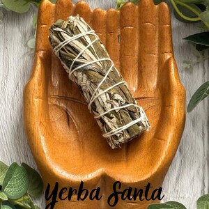May include: A wooden dish carved into the shape of two cupped hands holding a bundle of dried Yerba Santa leaves wrapped in white string. The wood has a warm brown tone with visible grain, and the text Yerba Santa is printed on the surface.
