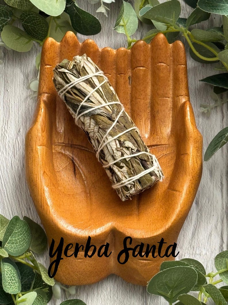 May include: A wooden dish carved into the shape of two cupped hands holding a bundle of dried Yerba Santa leaves wrapped in white string. The wood has a warm brown tone with visible grain, and the text Yerba Santa is printed on the surface.