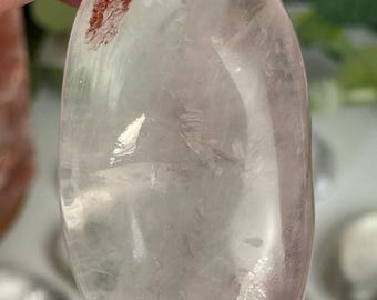 May include: A clear, oval-shaped crystal with a pale pink hue and a small, reddish-brown inclusion. The polished stone is held in a hand, showcasing its smooth surface and natural variations. The crystal is a natural gemstone.