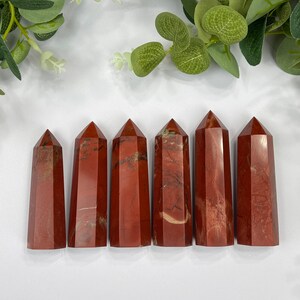 May include: Seven red jasper crystal points, each with a unique pattern of red and white, are arranged in a row on a white surface.