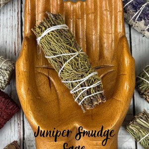 May include: A wooden bowl carved into the shape of two cupped hands holding a bundle of green juniper smudge sage wrapped in white string. The text Juniper Smudge Sage is printed in black script across the center of the wooden hand bowl.