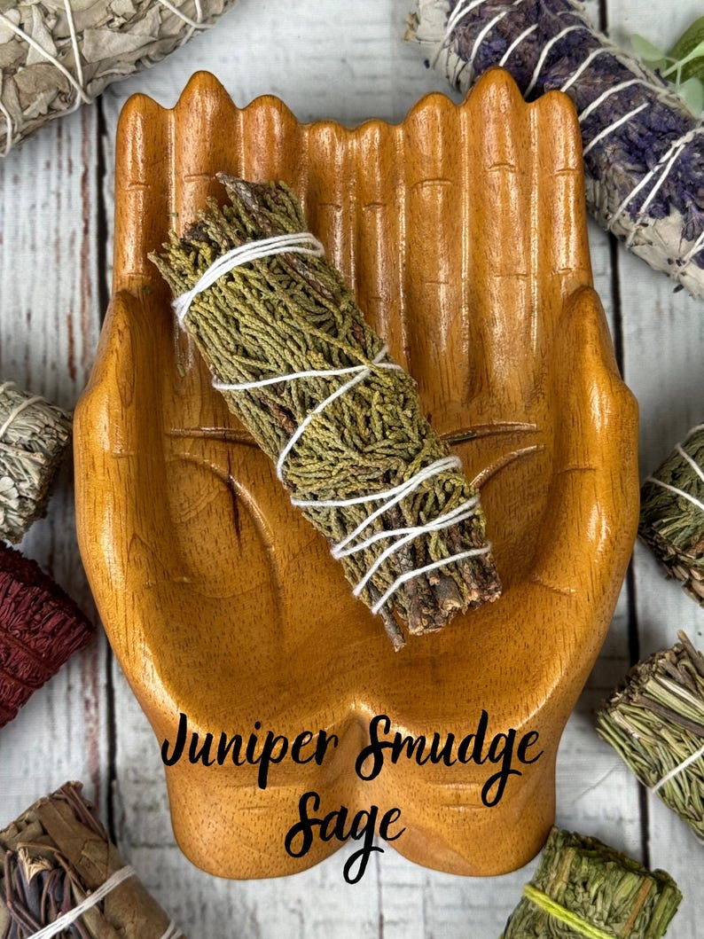 May include: A wooden bowl carved into the shape of two cupped hands holding a bundle of green juniper smudge sage wrapped in white string. The text Juniper Smudge Sage is printed in black script across the center of the wooden hand bowl.