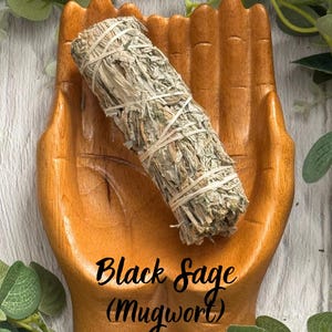 May include: A bundle of dried Black Sage (Mugwort) tied with white string, resting in a carved wooden dish shaped like two open hands. The sage is light green and brown, while the wooden dish has a smooth, warm, medium-brown finish.
