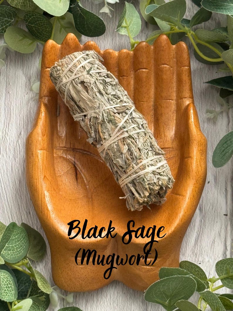 May include: A bundle of dried Black Sage (Mugwort) tied with white string, resting in a carved wooden dish shaped like two open hands. The sage is light green and brown, while the wooden dish has a smooth, warm, medium-brown finish.