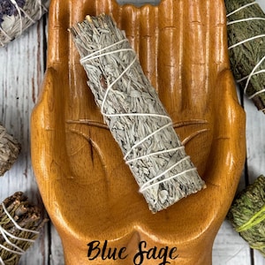 May include: A light brown wooden dish carved in the shape of two cupped hands holding a bundle of dried blue sage wrapped in white string. The text Blue Sage is printed in black script on the palm area of the wooden hand-shaped dish.
