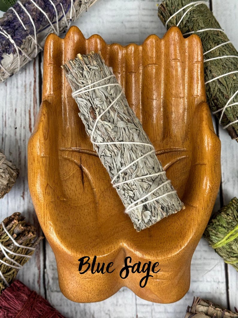 May include: A light brown wooden dish carved in the shape of two cupped hands holding a bundle of dried blue sage wrapped in white string. The text Blue Sage is printed in black script on the palm area of the wooden hand-shaped dish.