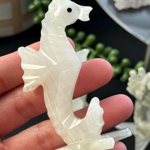 Puede incluir: Figurita de caballito de mar tallada en cristal de selenita blanco. El caballito de mar est&aacute; de pie sobre una peque&ntilde;a base y tiene un ojo negro.