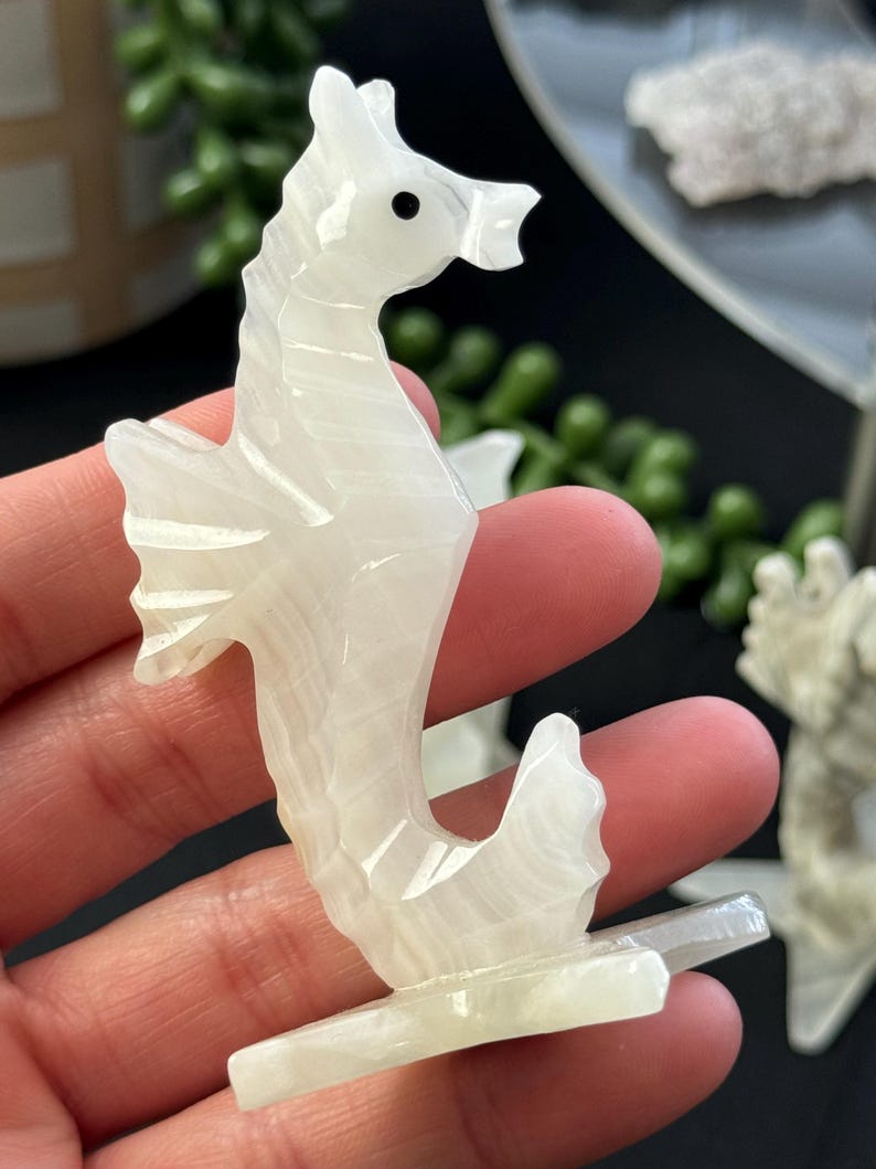 Puede incluir: Figurita de caballito de mar tallada en cristal de selenita blanco. El caballito de mar est&aacute; de pie sobre una peque&ntilde;a base y tiene un ojo negro.