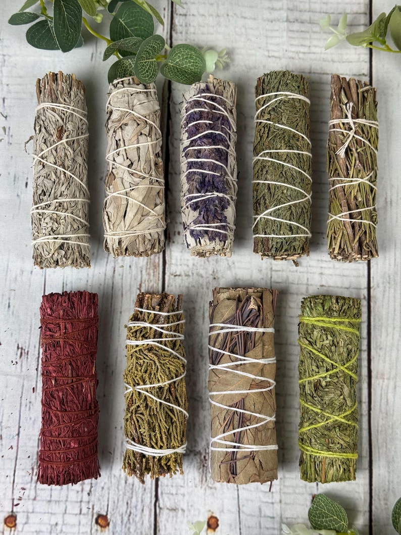 May include: A collection of nine cylindrical smudge sticks arranged in two rows. The bundles feature various dried herbs, leaves, and flowers in shades of beige, green, purple, and red, each tightly secured with thin white or colored string.