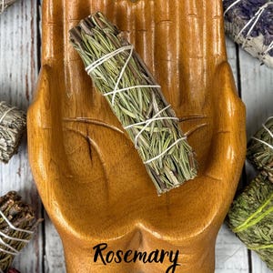 May include: A polished wooden dish shaped like two cupped hands, featuring the word Rosemary printed in black script at the base. A bundle of dried green rosemary stems tied with white string rests inside the center of the wooden hand dish.