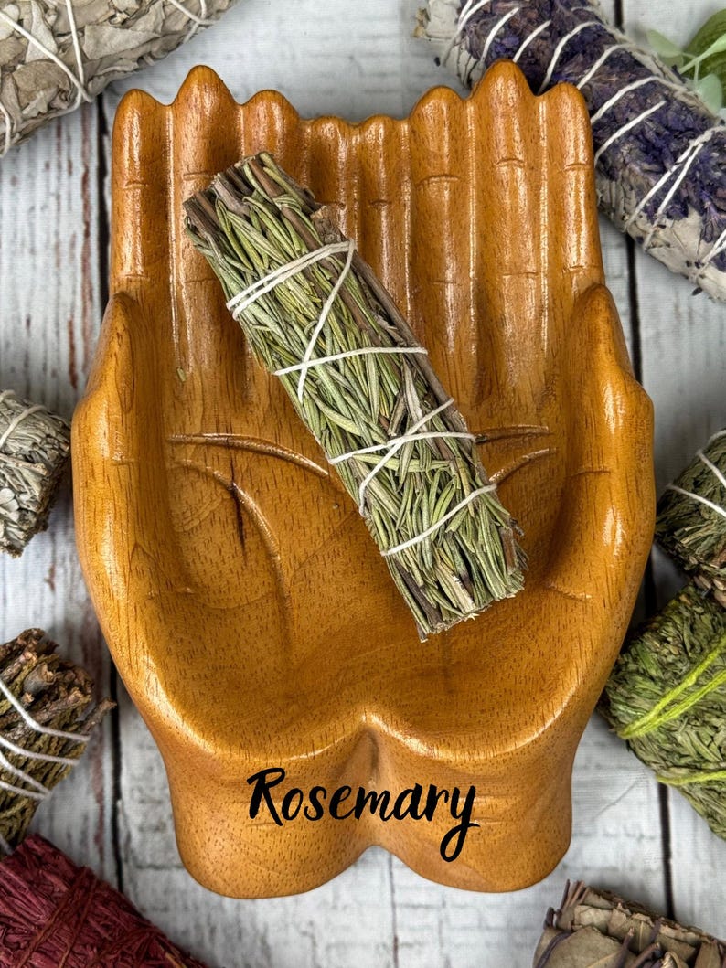 May include: A polished wooden dish shaped like two cupped hands, featuring the word Rosemary printed in black script at the base. A bundle of dried green rosemary stems tied with white string rests inside the center of the wooden hand dish.