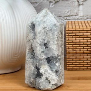 May include: A light blue celestite crystal tower with a faceted top. The crystal has a rough, textured surface and a hollowed-out interior. The crystal is displayed on a wooden surface.