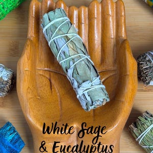 May include: A wooden dish shaped like two cupped hands holding a bundle of white sage and eucalyptus tied with white string. The text White Sage & Eucalyptus is printed in black on the palm of the wooden hand dish, surrounded by other herb bundles.