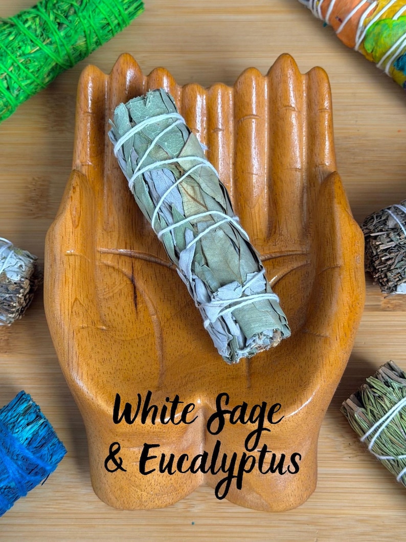 May include: A wooden dish shaped like two cupped hands holding a bundle of white sage and eucalyptus tied with white string. The text White Sage & Eucalyptus is printed in black on the palm of the wooden hand dish, surrounded by other herb bundles.
