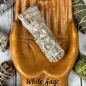 May include: A bundle of white sage wrapped in thin white string resting inside a carved wooden dish shaped like two cupped hands. The text White Sage is printed in black script at the bottom of the wooden dish, surrounded by other herb bundles.