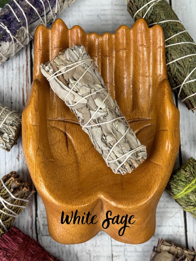 May include: A bundle of white sage wrapped in thin white string resting inside a carved wooden dish shaped like two cupped hands. The text White Sage is printed in black script at the bottom of the wooden dish, surrounded by other herb bundles.