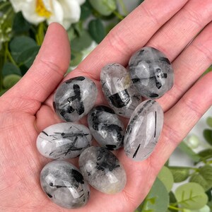May include: A collection of tumbled black tourmaline quartz stones. The stones are oval-shaped, with clear quartz and black tourmaline inclusions. The stones are held in a hand, with a blurred floral background.
