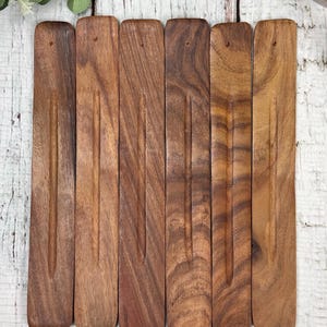 May include: Six brown wooden incense sticks with grooves carved into the surface. The sticks are arranged in a row on a white wooden surface.