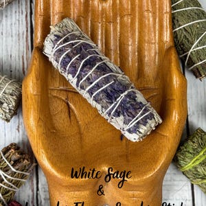 May include: A wooden hand-shaped dish holding a white sage and lavender flower smudge stick wrapped in white string. The text White Sage & Lavender Flower Smudge Stick is printed on the dish. Other smudge sticks are visible in the corners.