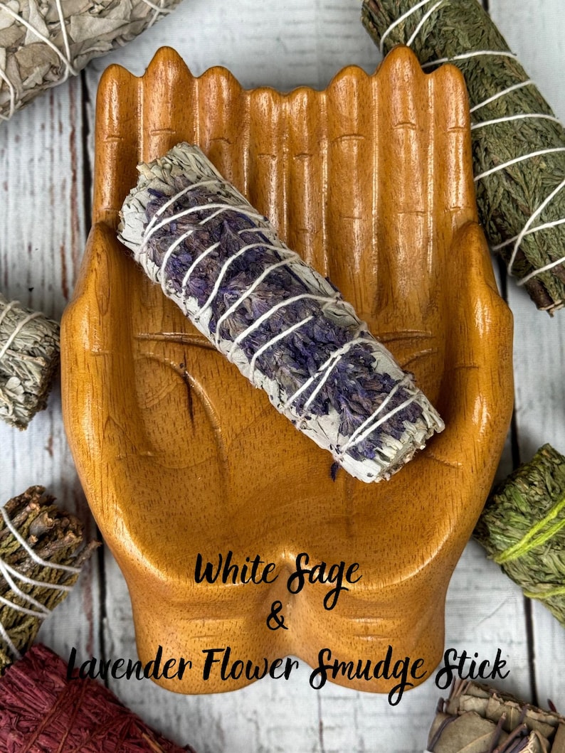 May include: A wooden hand-shaped dish holding a white sage and lavender flower smudge stick wrapped in white string. The text White Sage & Lavender Flower Smudge Stick is printed on the dish. Other smudge sticks are visible in the corners.