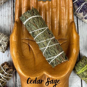 May include: A wooden dish shaped like two cupped hands holding a bundle of green cedar sage wrapped in white string. The text Cedar Sage is printed in black script on the palm of the wooden hand dish, which is surrounded by other herb bundles.