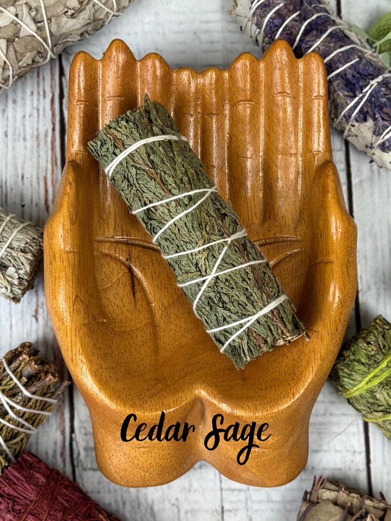May include: A wooden dish shaped like two cupped hands holding a bundle of green cedar sage wrapped in white string. The text Cedar Sage is printed in black script on the palm of the wooden hand dish, which is surrounded by other herb bundles.