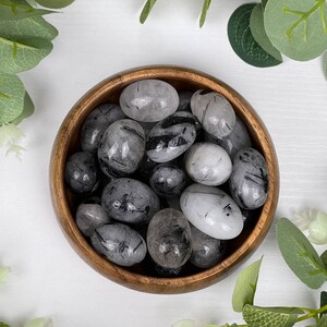 May include: A wooden bowl filled with polished black tourmaline stones. The stones are oval-shaped with a mix of black and translucent gray colors. Green leaves surround the bowl on a white surface.