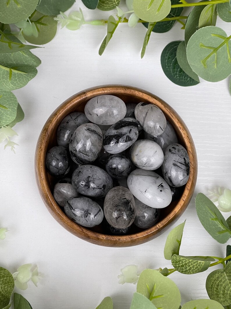May include: A wooden bowl filled with polished black tourmaline stones. The stones are oval-shaped with a mix of black and translucent gray colors. Green leaves surround the bowl on a white surface.