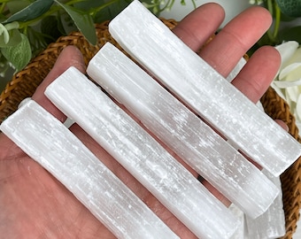 May include: Several white selenite crystal wands are displayed in a woven basket. The wands are rectangular and have a smooth, opaque appearance. The crystals are held in a hand, with greenery in the background.