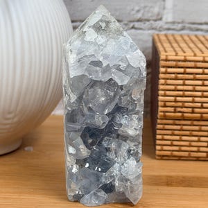 May include: A blue celestite crystal tower with a faceted, pointed top. The crystal is a light blue color with darker blue inclusions and sits on a wooden surface. A white vase and a woven basket are in the background.