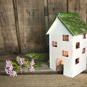 Könnte beinhalten: Weißes Keramikhaus mit einem grün glasierten Dach und ausgeschnittenen Fenstern. Das Haus ist ein Kerzenhalter mit einer kleinen Kerze im Inneren.