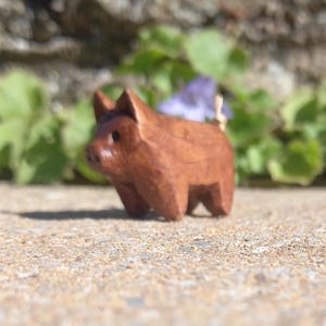 Puede incluir: Una pequeña figura de cerdo de madera tallada a mano. El cerdo es de color marrón cálido y tiene un diseño simple y en bloques. Se encuentra sobre una superficie texturizada, con follaje verde y una pared de piedra al fondo.
