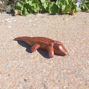 以下が含まれることがあります： 茶色の仕上げが施された、手彫りの木製ワニの置物。ワニは腹ばいになって口を閉じている。