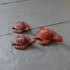 May include: Three wooden turtle figurines carved in a reddish-brown colour. The turtles are arranged in a row on a grey wooden surface.