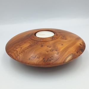 May include: A handcrafted wooden candle holder with a natural wood grain finish. The candle holder is oval-shaped with a circular indentation to hold a white candle. The wood has a warm, reddish-brown hue.